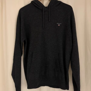 Mörkgrå kashmir hoodie från GANT - Mörkgrå hoodie från GANT i en kashmir blend med huva och snörning. Klassisk logga på bröstet och praktisk magficka framtill. Långärmad och perfekt för kyliga dagar. Snygg och enkel design som funkar till jeans eller mjukis. Nypris 2600!