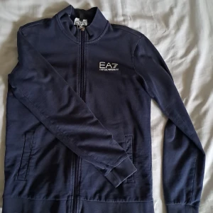 Marinblå ziptröja EA7 Emporio Armani - Snygg marinblå ziptröja från EA7 Emporio Armani med vit logga på bröstet. Tröjan har lång ärm, ståkrage och två fickor fram. Materialet är mjuk bomull och polyester, perfekt för chill eller sport. Dragkedja hela vägen framtill för enkel på- och avtagning.