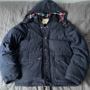 Mörkblå Jacka från  Abercrombie & Fitch - Säljer en riktigt sällsynt jacka från Abercrombie & Fitch i storlek S. Jackan har huva, dragkedja och knappar framtill samt två stora fickor. Insidan är fodrad med röd, blå och vit rutig bomull. Perfekt för kalla dagar. Ny pris 3000kr. Mitt pris 1499