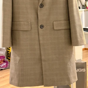beige rutig trenchcoat - Snygg beige rock från EPTISON med rutigt mönster och två knappar framtill. Jackan har klassisk krage och fickor med lock. Perfekt för dig som vill sticka ut med coola detaljer.
