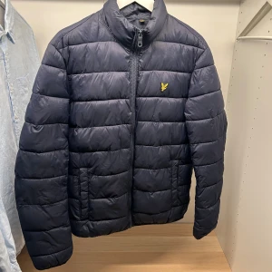 Mörkblå pufferjacka från Lyle & Scott - Snygg mörkblå pufferjacka från Lyle & Scott med gul logga på bröstet. Jackan har hög krage, dragkedja framtill och två sidofickor. Perfekt för kyliga dagar och har en klassisk quiltad look. Materialet är polyester och jackan har en normal passform.