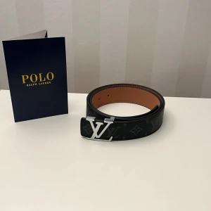  svart skärp  - Säljer ett stilrent svart bälte i skinn med klassiskt monogrammönster och ett stort, silverfärgat spänne. Insidan är brun med snygga vita sömmar. Perfekt accessoar för dig som vill ha en lyxig touch på outfiten.