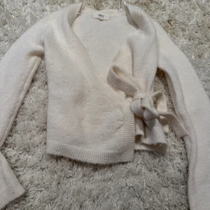 Vit omlottkofta med knyt från Gina tricot - Supermjuk vit omlottkofta från Monki med långa ärmar och breda ribbade muddar. Koftan har en snygg knytning i sidan och är stickad i ett fluffigt material som känns riktigt cozy. Perfekt för lager-på-lager och chill dagar.