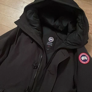 Canada Goose Chateau parka - Säljer en svart dunjacka från Canada Goose med stor huva, dragkedja och knappar framtill. Jackan har klassisk logga på ärmen och är riktigt varm och skön. Perfekt för kalla vinterdagar och har en clean, stilren look.