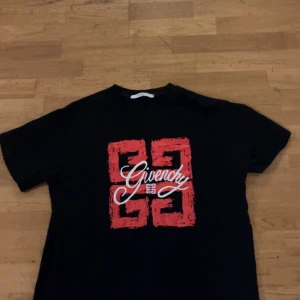 Svart Givenchy t-shirt med röd print - Svart t-shirt från Givenchy med ett stort rött grafiskt tryck och vit Givenchy-logga på bröstet. Klassisk passform och rund halsringning. Perfekt för dig som gillar streetwear och vill sticka ut med en exklusiv look.