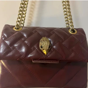 Kurt geiger väska - Snygg  axelväska i vintött skinn med guldfärgad kedjerem och unik fågeldekor på locket från Kurt Geiger . Väskan har ett klassiskt rutmönster och stängs med ett dekorativt spänne. Perfekt för dig som gillar detaljer och stilren design.