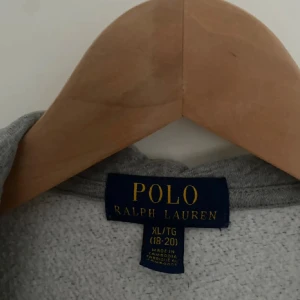 Grå hoodie från Polo Ralph Lauren XL - Snygg grå hoodie från Polo Ralph Lauren i storlek XL. Tröjan har dragkedja, huva och klassisk broderad logga på bröstet. Tillverkad i mjuk bomull och har kängurufickor framtill. Perfekt för chill dagar eller när du vill ha en clean look.