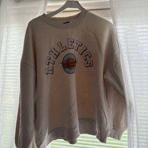 Beige sweatshirt - Säljer en oversized beige sweatshirt med trycket 'ATHLETICS Madison Wisconsin United States' i blått, vitt och rött framtill. Tröjan har rund hals, ribbade muddar och långa ärmar. Perfekt för en chill och sportig look. Från Gina Tricot!