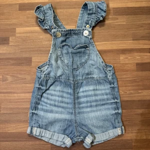 Hängselshorts  - Söta blå hängselshorts från H&M i storlek 68 (4-6mån). Jeansimitation, väldigt mjukt och skönt material. Volangdetaljer på axelband, knappar i metall och uppvikta benslut. Helt oanvänd, endast upptvättad.