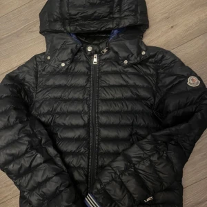 Svart Moncler dunjacka med huva - Svart dunjacka från Moncler med klassisk quiltad design, avtagbar huva och dragkedja framtill. Jackan har Moncler-logga på ärmen och ribbade muddar med vita ränder. Perfekt för kalla dagar och riktigt snygg streetstil.