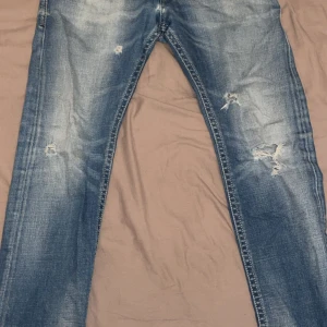 Ljusblåa Diesel jeans - Snygga blå jeans med slitningar och hål på framsidan för en cool, avslappnad look. Jeansen har raka ben och klassisk femficksdesign. Materialet är robust denim och färgen är tvättad blå med ljusare partier.