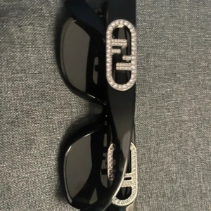 Svarta Fendi solglasögon med strass - Snygga svarta solglasögon från Fendi med rektangulära bågar och ikoniska Fendi-loggor i silver och strass på sidorna. Glasögonen har en lyxig känsla och passar perfekt för dig som vill sticka ut med en exklusiv accessoar. Pris går att diskutera