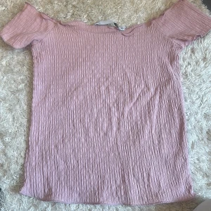 Ljusrosa offshoulder topp från 157 - Säljer en ljusrosa offshoulder topp från 157 i storlek XS. Toppen har en crinklad, ribbad struktur och är tillverkad i ett mjukt syntetmaterial. Den är ärmlös med breda axelpartier och har en slim passform som sitter snyggt mot kroppen.