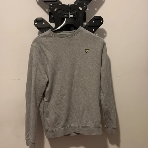 Grå sweatshirt från Lyle & Scott - Grå sweatshirt från Lyle & Scott med klassisk rund hals och långärmad design. Tröjan har en liten gul broderad logga på bröstet och är tillverkad i mjuk bomull. Perfekt för dig som gillar enkel och stilren look.
