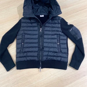 Svart pufferjacka från Moncler - Svart pufferjacka från Moncler med stickade ärmar och huva. Jackan har ribbade muddar, två fickor med knapp framtill och en ficka med dragkedja på ärmen. Materialmix av polyester och stickad ull ger en schysst look. Perfekt för dig som vill ha något både snyggt och funktionellt.