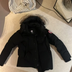 Svart Canada Goose dunjacka med päls - Svart dunjacka från Canada Goose med ikonisk patch på ärmen och fluffig päls runt huvan. Jackan har dragkedja och knappar framtill, långa ärmar och flera fickor. Perfekt för kalla vinterdagar och har en clean, klassisk look.