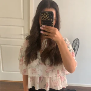 Blommig volangblus med knyt - Säljer en söt blommig blus i vitt med rosa och gröna blommor. Blusen har volanger, v-ringning och knyt framtill vid halsen. Den är luftig och har korta ärmar med volangdetaljer. Perfekt för en somrig look! Slutsåld ord pris runt 400kr