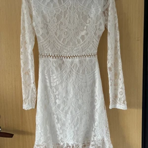 Vit spetsklänning med öppen rygg - Superfin vit kort spetsklänning med lång ärm och snygg öppen rygg. Klänningen har vackra spetsdetaljer, markerad midja och knyt i nacken. Använder ej för de är inte min stil 