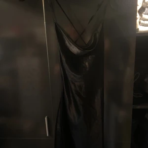Svart långklänning med tunna band - Säljer en svart långklänning med tunna axelband som korsas i ryggen. Klänningen har en snygg, glansig finish och ett elegant fall. Perfekt för dig som gillar minimalistisk och stilren look. Den går ungefär ner till knäna