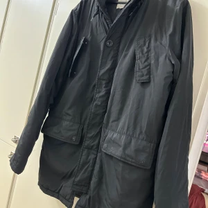 Svart parkajacka med dragkedja - Svart parkajacka  med dragkedja och knappar framtill. Jackan har flera stora fickor med lock och en hög krage. Materialet är syntet och jackan är helfodrad för extra värme. Perfekt för kyliga dagar och har en stilren, enkel look.