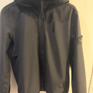 Svart softshelljacka Stone Island - Svart softshelljacka från Stone Island med huva och dragkedja framtill. Jackan har klassisk patch på ärmen och är tillverkad i stretchig polyester med vattenavvisande och vindtät teknologi. Perfekt för dig som vill ha en clean och stilren look.