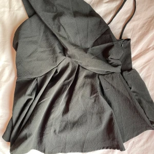 Svart topp - Snygg svart topp med asymmetrisk design och en axelbandslös sida samt ett smalt axelband på andra sidan. Toppen har ett lätt rynkat tyg och markerad midja som ger en cool och modern siluett. Perfekt för dig som gillar unika detaljer och stilren look.