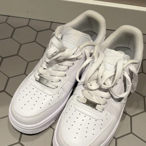 Nike Air Force 1 vita sneakers - Fräscha Nike Air Force 1 sneakers i helvitt med klassisk design. Skorna har snörning, perforerad tå och platt sula. Tillverkade i skinn med mjuk textil på insidan. Perfekta för dig som gillar stilrena och tidlösa sneakers. Endast testade inomhus, men för små.