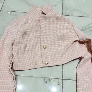 Croppad rosa rutig kavaj med pärlknappar - Säljer en croppad kavaj i ljusrosa och vitt med rutigt mönster. Jackan har högt ståkrage, långa ärmar och två snygga pärlknappar med gulddetaljer framtill. Materialet är strukturerat och känns lyxigt, perfekt för att lyfta din outfit.