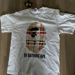 Vit t-shirt från A Bathing ape ( Bape) - Vit t-shirt från Bape med klassisk rund hals och korta ärmar. Stor apansikte-tryck på framsidan med rutigt mönster i beige, rött och svart. Materialet är mjuk bomull och etiketten visar tydligt BAPE-loggan. Perfekt för dig som gillar stil 