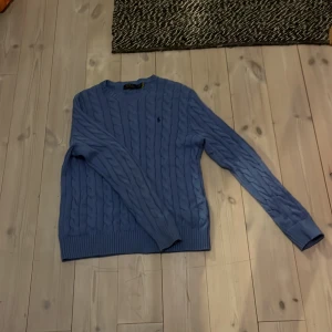 Blå kabelstickad tröja Polo Ralph Lauren - Snygg blå kabelstickad tröja från Polo Ralph Lauren i storlek M. Tröjan har rund hals, långärmade ribbade muddar och klassisk broderad logga på bröstet. Perfekt för dig som gillar stilrena och tidlösa plagg med premiumkänsla.