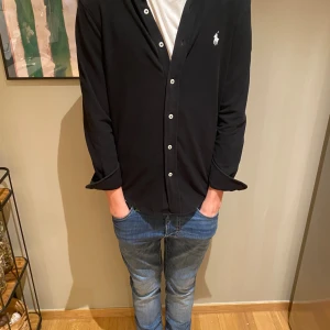 Svart skjorta från Polo Ralph Lauren XS - Svart långärmad skjorta från Polo Ralph Lauren i lätt mesh-bomull. Klassisk button-down krage, vita knappar och ikonisk broderad logga på bröstet. Perfekt för dig som gillar stilrena och tidlösa plagg.