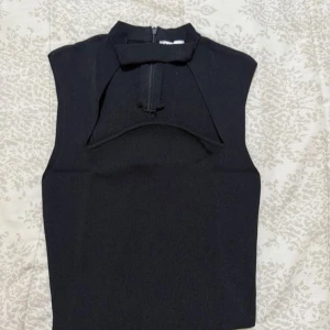 Svart  ärmlös polotopp med dragkedja - Snygg svart polotopp med hög krage och cool dragkedjedetalj framtill. Toppen är ärmlös och har en stilren, tight passform som ger en modern vibe. Materialet är ribbat och känns mjukt mot huden. Perfekt för dig som gillar minimalistisk och edgy stil.