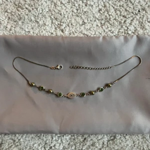 Silverfärgat halsband med gröna pärlor/stenar - Unikt halsband i silver med små gröna pärlor , stenar och ett öga i mitten. Kedjan är tunn och har justerbar längd med karbinlås. Perfekt för dig som vill ha något stilrent men ändå med en cool detalj. Som längst 41cm och minst 36cm 