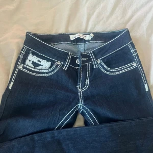 Mörkblå My Christy's jeans med broderi - Snygga lågmidjade mörkblå jeans från My Christy's med vit broderad söm och detaljerade fickor bak med knappar. Jeansen har klassisk design och markerade sömmar som ger en cool och unik look. Säljer då dom är för små för mig och inte längre kommer till användning!💓