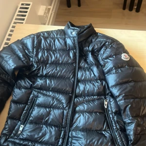 Svart dunjacka från Moncler - Snygg svart dunjacka från Moncler med glansig finish och klassisk quiltad design. Jackan har ståkrage, två dragkedjefickor framtill och Moncler-logga på ärmen. Perfekt för kalla vinterdagar och riktigt trendig stil.