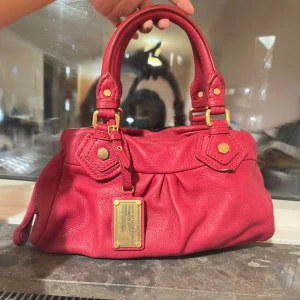 Röd handväska från Marc by Marc Jacobs - Cool röd handväska i mjukt skinn från Marc by Marc Jacobs med guldfärgade metalldetaljer och dekorativ loggoplatta framtill. Väskan har dubbla handtag, dragkedja upptill och snygga veckade detaljer på framsidan.