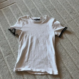 Vit t-shirt med spetsdetalj från Zara - Vit t-shirt från Zara med korta ärmar och svart spetsdetalj med pärlor vid ärmsluten. T-shirten har en klassisk rund hals och är gjord i mjuk bomull. Passformen är normal och modellen är enkel men med en cool twist på ärmarna.