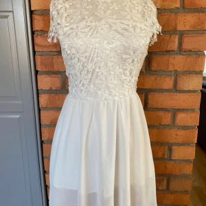 Vit spetsklänning med blommönster strl M - Superfin vit klänning med blommig spets upptill och korta ärmar. Kjolen är lätt och flowy, medan överdelen har ett vackert broderat blommönster. Dragkedja i ryggen för enkel påklädning. Från Nilanki Fashion