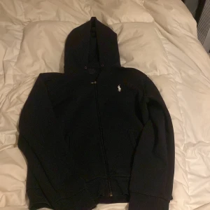 Svart hoodie från Ralph Lauren - Svart hoodie från Ralph Lauren med dragkedja och huva. Klassisk design med den vita Polo-loggan broderad på bröstet. Perfekt för chill dagar och enkel att matcha med jeans eller joggers.