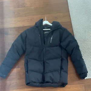 Vinter jacka sail Racing - Svart pufferjacka från Sail Racing med huva och dragkedja framtill. Jackan har en stilren design med logga på bröstet och är perfekt för kalla dagar. Fylld för extra värme och har praktiska fickor med dragkedja.