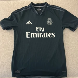 Real Madrid svart matchtröja Adidas M - Svart Real Madrid fotbollströja från Adidas, säsong 18/19 med Fly Emirates-tryck på bröstet. Kortärmad med diskreta ränder och klubbmärke på vänster sida. Tillverkad i lätt och ventilerande Climachill-material, perfekt för träning eller match.