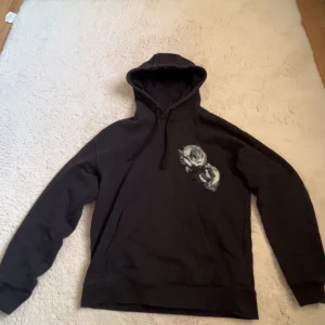 Svart hoodie med rosor från HUGO - Svart hoodie från HUGO med snyggt tryck av vita och grå rosor på både fram- och baksidan. Stor HUGO-logga på ryggen, huva med snörning och klassisk känguruficka. Perfekt för dig som gillar streetstyle och vill sticka ut lite extra.