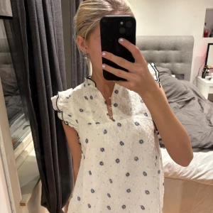 Vit blommig blus från Lindex - Supersöt vit blus från Lindex med mörkblå blommigt mönster och små ljusblå detaljer. Blusen har volangärmar med mörkblå kant, v-ringad hals med knappar och lätt struktur i tyget. Perfekt för dig som gillar romantisk och fräsch stil.