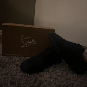 Svarta sneakers från Christian Louboutin - Snygga svarta sneakers från Christian Louboutin med klassisk röd sula. Skorna har rund tå, snörning och är tillverkade i mocka och skinn. Perfekta för dig som vill ha en stilren och exklusiv look med en ikonisk touch.