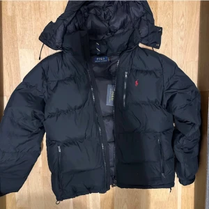Svart dunjacka Polo Ralph Lauren - Svart dunjacka från Polo Ralph Lauren med avtagbar huva, dragkedjor på fickor och bröst, samt röd broderad logga på bröstet. Jackan är quiltad och har en klassisk pufferstil som håller dig varm under kalla dagar.