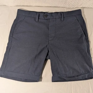 Mörkblå shorts från Jack & Jones - Snygga mörkblå shorts från Jack & Jones med klassisk passform. De har två sidofickor, två bakfickor med knapp och hällor för bälte. Materialet känns mjukt och svalt, perfekt för varma dagar. Stängs med knapp och dragkedja framtill.