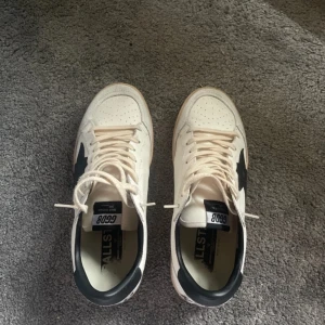 Golden Goose Ball Star sneakers vit/svart - Golden Goose Ball Star sneakers i vitt skinn med svart stjärndetalj på sidan och svart hälkappa. Klassisk låg modell med snörning, perforerad tå och texten 'SNEAKERS' på hälen. Snygg retrovibe och platt sula, perfekt till jeans eller shorts.