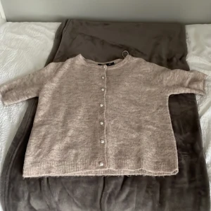 Beige stickad kofta med pärlknappar - Mysig beige stickad kofta från Vero Moda med pärlknappar framtill. Koftan har rund hals, trekvartsärmar och ribbade muddar vid nederkant och ärmslut. Perfekt för lager-på-lager och chill dagar. Snygg att matcha med jeans eller kjol.