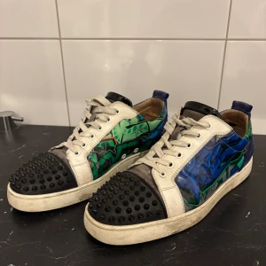 Färgglada sneakers med nitar - Unika sneakers med coolt grafiskt mönster i grönt och blått på sidorna, vita partier och svarta tåhättor med nitar. Skorna har snörning, rund tå och detaljer i skinn. Perfekta för dig som vill sticka ut med din stil.