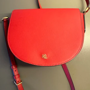 Röd axelväska från Ralph Lauren - Snygg röd axelväska från Ralph Lauren med guldfärgad RLL-logga framtill. Väskan har en rundad form och justerbar axelrem med gulddetaljer. Tillverkad i slätt skinn som ger en lyxig känsla. Perfekt storlek för det viktigaste och stilren design som lyfter din outfit.